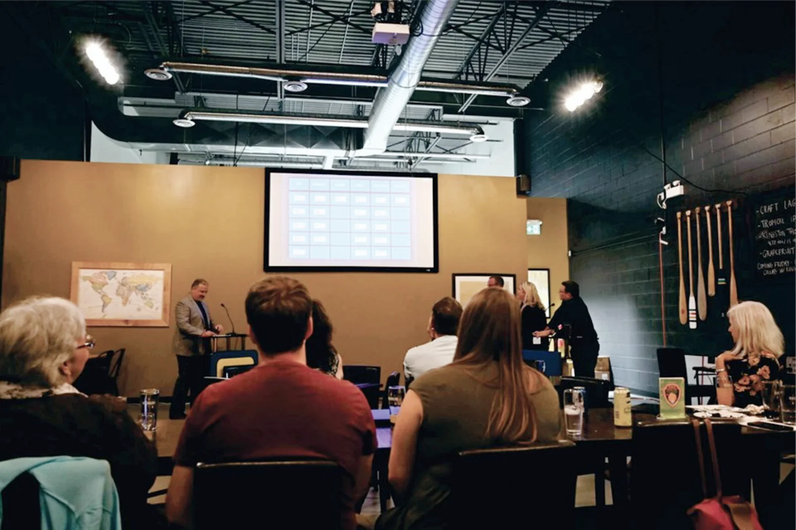 A group of attendees in a conference room are engaged in a presentation, with a speaker at the podium in front of a projection screen displaying a grid layout. The setting features a map on the wall, accent lighting, and decorative paddles, creating a collaborative atmosphere for the event.