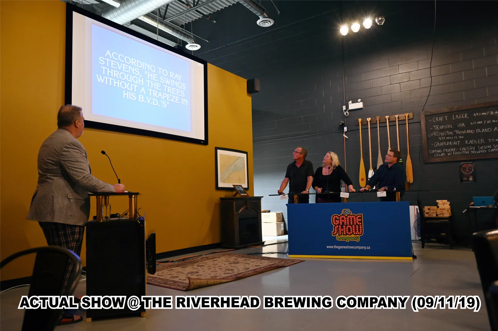Image of a live trivia game show event at The Riverhead Brewing Company on September 11, 2019. A host stands at a podium, with a large screen displaying a quote from Ray Stevens. Three contestants are engaged, standing at a game show desk. The venue has a casual atmosphere, featuring decor and seating arrangements.