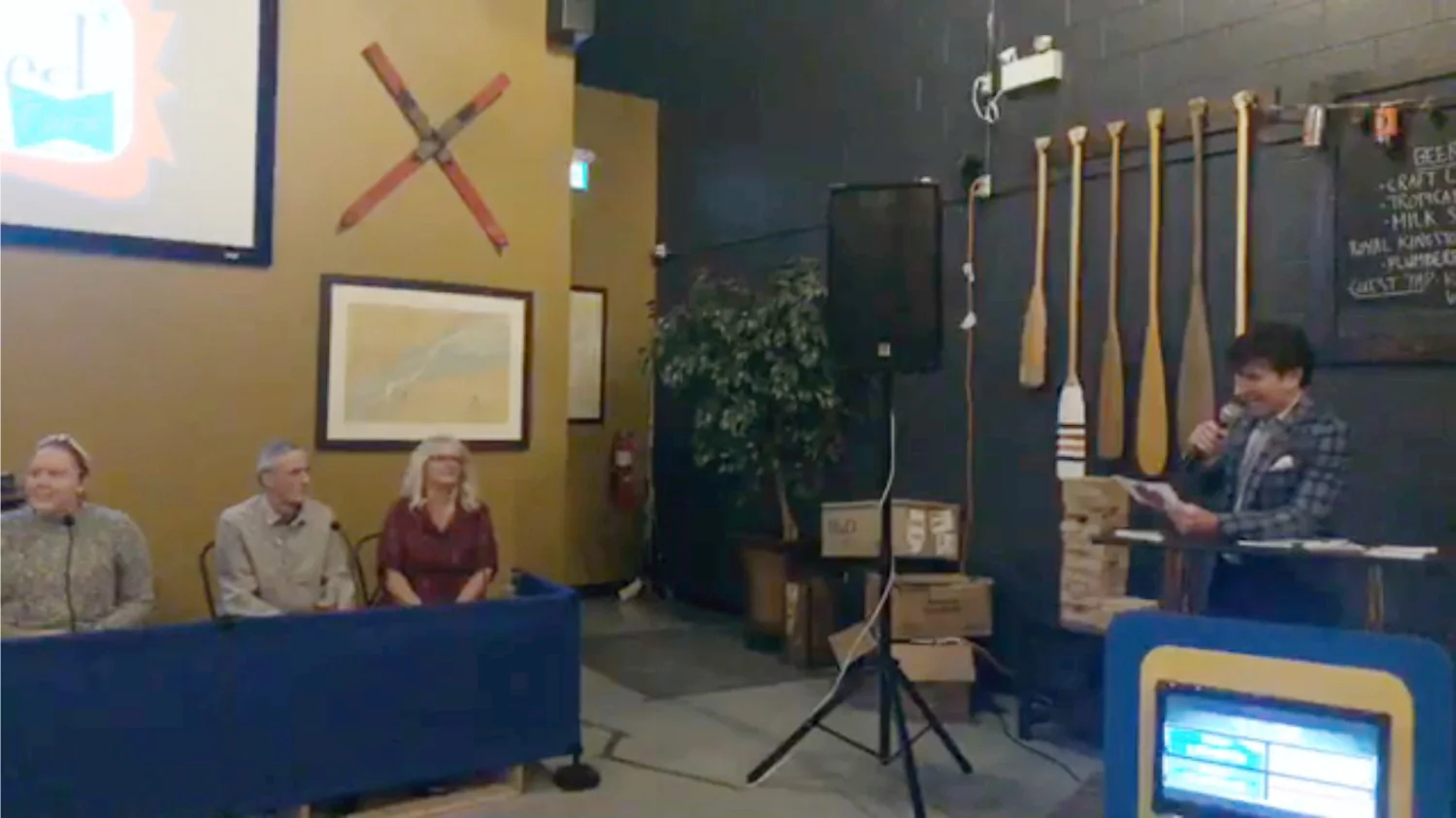 Panel discussion event featuring four speakers seated at a table, with a projector displaying a logo on a wall. The setting includes decorative paddles and artwork, contributing to a cozy atmosphere. A microphone is held by the moderator at the forefront, engaging with the audience.