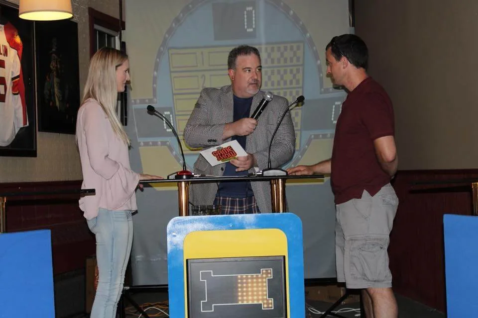 Two participants engage in a quiz game on stage while a host presents questions. The backdrop features a large game board, and the setting appears to be a lively venue with sports memorabilia on display. The audience is likely entertained by the interactive game show experience.