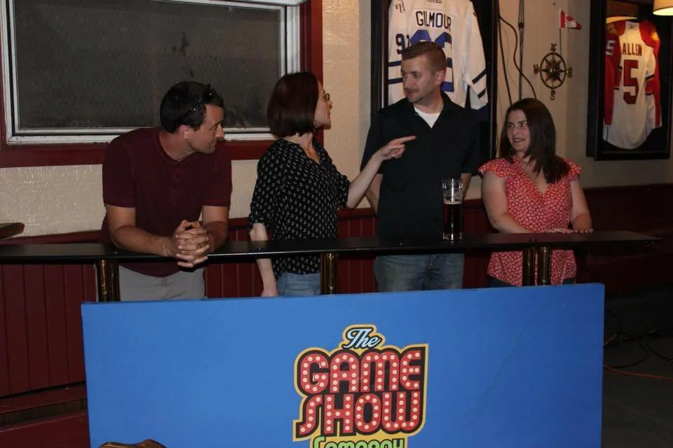 Group of four individuals engaged in a lively conversation at a game show-themed event, with a blue sign reading "The Game Show" in front. The setting features a bar environment with sports memorabilia displayed on the walls.
