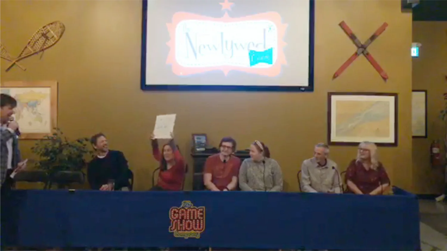 Participants at a live game show event engage in the Newlywed Game, with one contestant holding up a card while others sit at the table, showcasing excitement and interaction. A screen in the background displays the game show title, adding to the lively atmosphere.