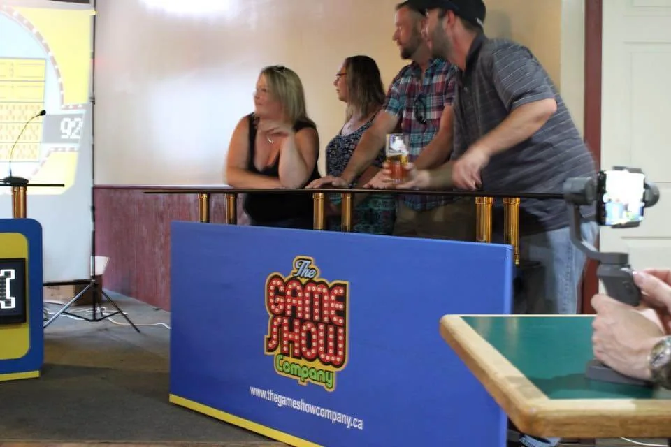 Group of participants engaging in a live game show event, with a colorful game show backdrop and a digital scoreboard visible. Some participants are cheering and one is holding a drink, while others appear to be focused on the game. The setting includes game show equipment in a lively atmosphere.
