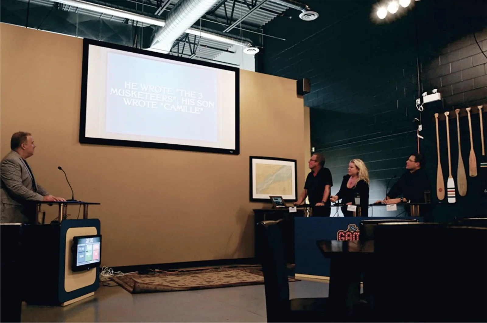A speaker presents to an audience at a literary event, discussing famous works such as "The Three Musketeers" and "Camille." The backdrop features a screen displaying text related to the presentation, while audience members engage attentively.